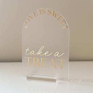 Puede incluir: Letrero de acr&iacute;lico transparente con la parte superior arqueada y una base rectangular. El letrero tiene letras doradas que dicen "LOVE IS SWEET" en la parte superior y "take a TREAT" en el centro. Dise&ntilde;ado para una boda o fiesta.