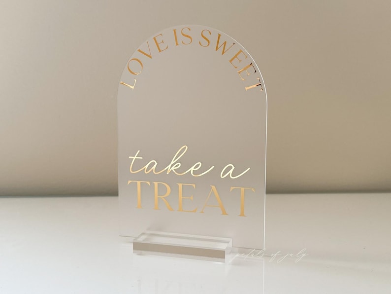 Puede incluir: Letrero de acr&iacute;lico transparente con la parte superior arqueada y una base rectangular. El letrero tiene letras doradas que dicen "LOVE IS SWEET" en la parte superior y "take a TREAT" en el centro. Dise&ntilde;ado para una boda o fiesta.