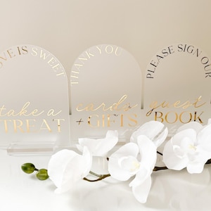 Puede incluir: Tres letreros de boda de acr&iacute;lico transparente con letras doradas. Los letreros dicen "Love is Sweet, take a Treat", "Thank You, cards + gifts" y "Please sign our guest book". Orqu&iacute;deas blancas en primer plano.