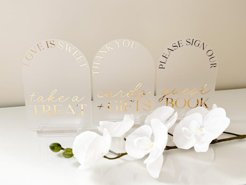 Puede incluir: Tres letreros de boda de acr&iacute;lico transparente con letras doradas. Los letreros dicen "Love is Sweet, take a Treat", "Thank You, cards + gifts" y "Please sign our guest book". Orqu&iacute;deas blancas en primer plano.