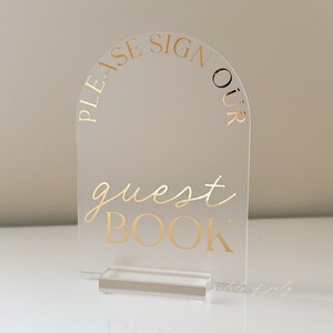 Puede incluir: Letrero de libro de visitas de acr&iacute;lico transparente con letras doradas. El letrero tiene forma de arco en la parte superior y dice "PLEASE SIGN OUR GUEST BOOK" en escritura elegante y may&uacute;sculas. El letrero se sujeta con una base de acr&iacute;lico transparente.