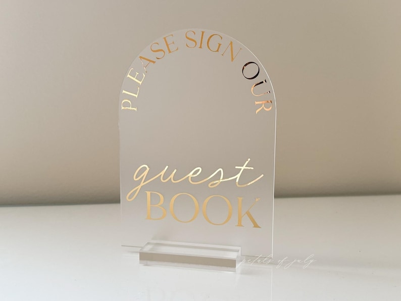 Puede incluir: Letrero de libro de visitas de acr&iacute;lico transparente con letras doradas. El letrero tiene forma de arco en la parte superior y dice "PLEASE SIGN OUR GUEST BOOK" en escritura elegante y may&uacute;sculas. El letrero se sujeta con una base de acr&iacute;lico transparente.