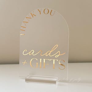 Puede incluir: Letrero de acr&iacute;lico transparente con letras doradas. El texto dice "THANK YOU" en la parte superior y "cards + GIFTS" debajo. El letrero tiene una parte superior arqueada y se encuentra en una base de acr&iacute;lico transparente. Ideal para bodas o eventos.