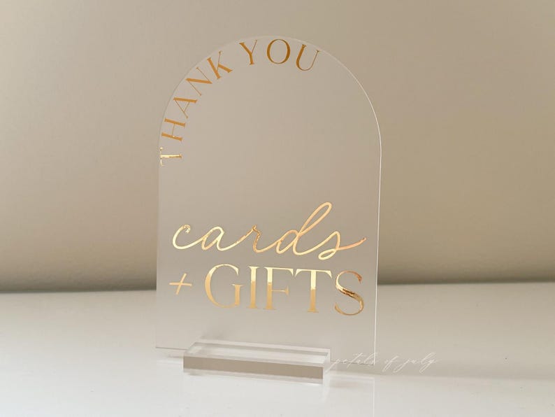 Puede incluir: Letrero de acr&iacute;lico transparente con letras doradas. El texto dice "THANK YOU" en la parte superior y "cards + GIFTS" debajo. El letrero tiene una parte superior arqueada y se encuentra en una base de acr&iacute;lico transparente. Ideal para bodas o eventos.