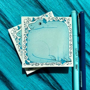 May include: Two square, light blue sticky notes with a cartoon dragon illustration and a decorative border. Two pens, one light blue and one dark blue, are next to the notes. The background is a textured, teal fabric.