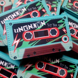 May include: A collection of teal cassette tape-themed wallets. Each wallet features the word "UNKNOWN" in white, red, and black, along with the text "TILL THE END" and "COME ON!". The design includes a red cassette tape graphic and a lightning bolt.