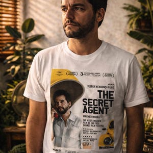 May include: A white t-shirt featuring a movie poster for "The Secret Agent." The poster includes the film's title, cast, and awards. The shirt is worn by a person with dark hair and a beard.