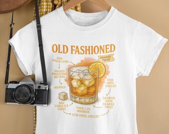 May include: White t-shirt with an "Old Fashioned" cocktail recipe graphic. The illustration shows a glass with ice cubes, an orange slice, and instructions for making the drink. The text includes "Add Sugar Cube", "Dashes Angostura Bitters", and "Pour 2 oz Bourbon".