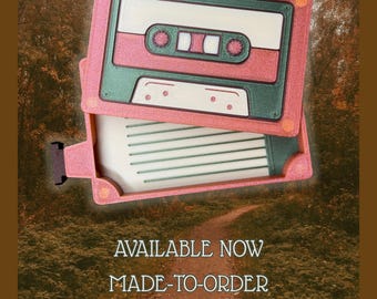 Op de afbeelding: Een rechthoekige opbergdoos in de vorm van een vintage cassettebandje met een roze frame, donkergroene accenten en een witte binnenkant. Het deksel heeft een cassetteontwerp met twee ronde openingen en een centraal venster.