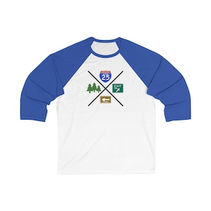 May include: White and blue baseball tee with a graphic of road signs, including "Interstate 25" and an exit sign. The design also features trees and a sign indicating 3 miles. The shirt has blue sleeves.