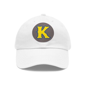 May include: White baseball cap with a gray circle patch featuring a yellow letter K with a blue outline.
