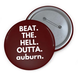 May include: A round, maroon button with the text "BEAT. THE. HELL. OUTTA. auburn." in white, with a silver pin back. The button is slightly angled, revealing the silver back of another button.