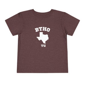 May include: A maroon toddler t-shirt with the white text "BTHO" above a white outline of the state of Texas, and the white text "TU" below the state outline. The t-shirt is made of a soft material.