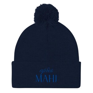 May include: A navy blue beanie with a matching pom-pom on top. The word "après" is embroidered in a cursive font above the word "MAHJ" in a block font. The beanie is a winter accessory.