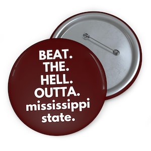 May include: A maroon button with the text "BEAT. THE. HELL. OUTTA. mississippi state." in white. The button has a silver pin back and is slightly angled, revealing the back of the button.