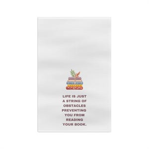 May include: A white tea towel with a stack of colorful books and a green leaf on top. The text "Life is just a string of obstacles preventing you from reading your book." is printed below the books.