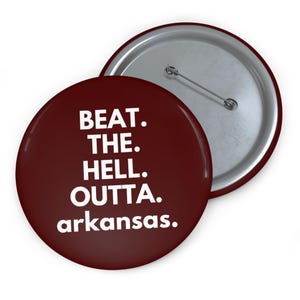 May include: A round, maroon button with the text "BEAT. THE. HELL. OUTTA. arkansas." in white, sans-serif font. The button has a silver pin back and a shiny, glossy finish. The button is a novelty item.