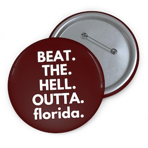 May include: A maroon button with the text "BEAT. THE. HELL. OUTTA. florida." in white. The button has a silver pin back and is shown next to another button.