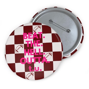 May include: A round button with a checkerboard pattern in maroon and white. The button features the text "BEAT. THE. HELL. OUTTA. t.u." in pink, along with football illustrations. The button has a metal pin back.