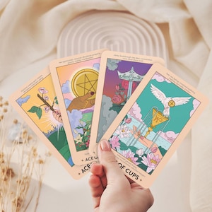 May include: Four tarot cards are fanned out, each featuring colorful illustrations. The cards depict images of a hand holding a coin, a dove above a chalice, and other symbolic scenes. The cards are held against a soft, neutral background.