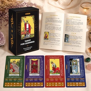 May include: A black tarot deck box labeled Tarot for Learning, an open guidebook, and four tarot cards: The Empress in green, Justice in purple, Strength in red, and The High Priestess in blue. Each card features illustrations and text.
