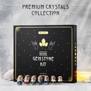 May include: A black box set labeled "Premium Crystals Collection" and "Natural Gemstone Kit." The box features gold accents and a graphic of hands holding crystals. Small glass bottles with cork stoppers containing colorful gemstones are in front of the box.