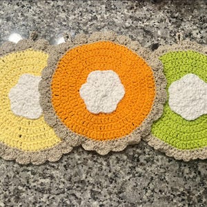 May include: Three handmade crochet pot holders in yellow, orange, and green. Each circular pot holder has a white flower-shaped center and a scalloped edge. The pot holders are approximately 7 inches in diameter.