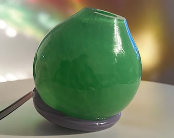 May include: A green ceramic vase with a rounded shape and a smooth, glossy finish. The vase is sitting on a purple base.