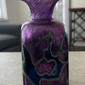 May include: A small, purple glass vase with a wide, flared opening and a square body. The vase has a swirling, iridescent pattern and black and blue accents.