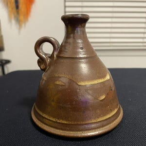 May include: A small, brown ceramic jug with a handle. The jug has a glazed finish and a light brown stripe around the middle.