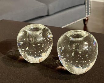 May include: Two clear, spherical glass candle holders with a bubble-like texture. Each holder has a circular indentation at the top for a candle. The candle holders are displayed on a dark brown surface.