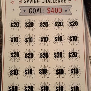 May include: A rectangular July saving challenge card with a $400 goal. The card features a US flag, firework-themed icons containing dollar amounts of $10, $20, and $50, and patriotic illustrations of two firework rockets and a star-shaped flag.