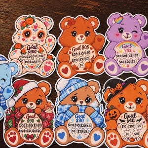 May include: Six die-cut stickers shaped like cartoon bears, each featuring a circular belly area with a savings goal amount and smaller circles labeled with dollar values. The bears have seasonal themes including floral, autumn, winter, and Halloween designs.