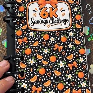 May include: A black binder insert featuring a repeating pattern of oranges, orange slices, white blossoms, and orange bows. The center displays a white rectangular label with an orange bow and the text 6K Savings Challenge in black font.