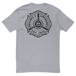 Può includere: Una t-shirt color grigio melange con una grafica nera e bianca invecchiata di un'elica e il testo "Engines & Airframes Est. 2023".