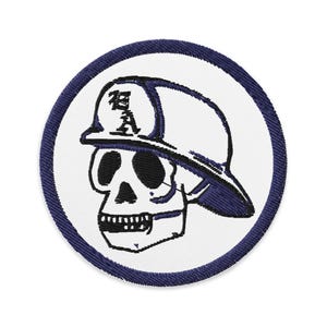 Può includere: Una toppa ricamata bianca con un bordo blu scuro, che presenta un teschio dei cartoni animati che indossa un berretto da baseball bianco e nero con la lettera "R" sul davanti.