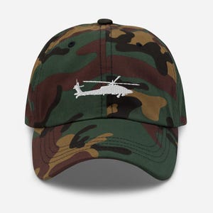 May include: Camouflage baseball cap with a white embroidered helicopter design. The cap features a curved brim and an adjustable strap for a comfortable fit. The hat's pattern includes shades of green, brown, and tan.
