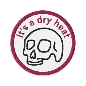 May include: A white embroidered patch with a pink border. The patch features a skull and the text "It's a dry heat".