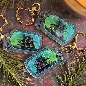 Peut inclure: Porte-clés en acrylique transparent en forme de bouteilles, chacun contenant une illustration de navire noir sur un fond vert et bleu. Les porte-clés ont une quincaillerie dorée et sont décorés d'étoiles et d'une ancre.