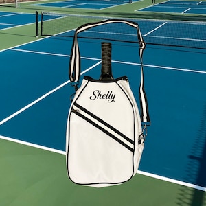 Puede incluir: Bolsa blanca para raqueta de tenis con detalles negros y una correa negra. La bolsa tiene el nombre "Shelly" bordado en la parte delantera.