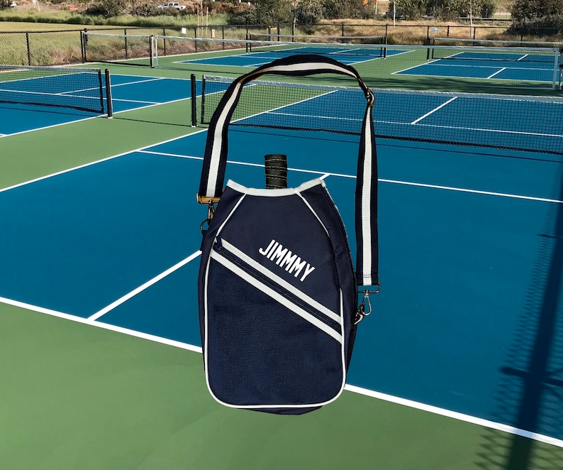 May include: A navy blue tennis racket bag with a white strap and white accents. The bag has the name "JIMMY" embroidered on the front.
