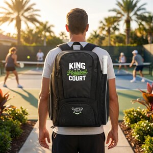 May include: A black backpack with a white and green graphic that reads KING OF THE PICKLEBALL COURT and the text CLAYCO SPORTS with a chevron logo. The backpack features a side pocket holding a white pickleball paddle handle.