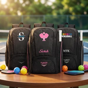 Op de afbeelding: Drie zwarte rugzakken ontworpen voor pickleball-uitrusting, elk met unieke personalisatie. De linker tas toont een grote letter S met de naam Smith. De middelste tas toont roze pickleball-peddels en de naam Sarah. De rechter tas bevat de tekst Pickleball en MTK. Alle tassen zijn voorzien van het Clayco Sports-logo.