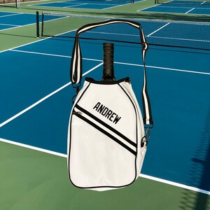 Puede incluir: Bolsa de raqueta de tenis blanca con una correa a rayas blancas y negras. La bolsa tiene una cremallera negra y el nombre "ANDREW" est&aacute; bordado en la parte delantera.