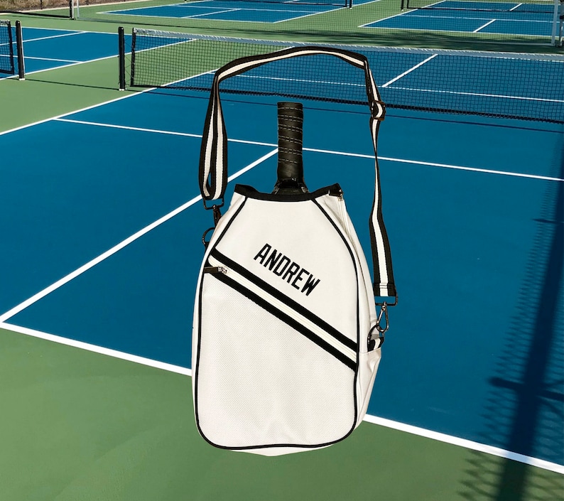 Puede incluir: Bolsa de raqueta de tenis blanca con una correa a rayas blancas y negras. La bolsa tiene una cremallera negra y el nombre "ANDREW" est&aacute; bordado en la parte delantera.
