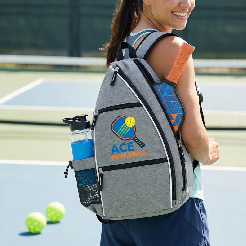 An adult woman is standing on a pickleball court, wearing a gray backpack with a pickleball paddle logo on it.