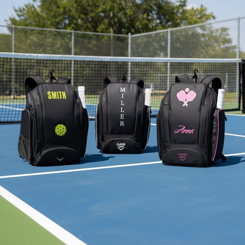 Three pickleball backpack bags with personalized names on them, placed on a pickleball court. One black, one white, and one pink.