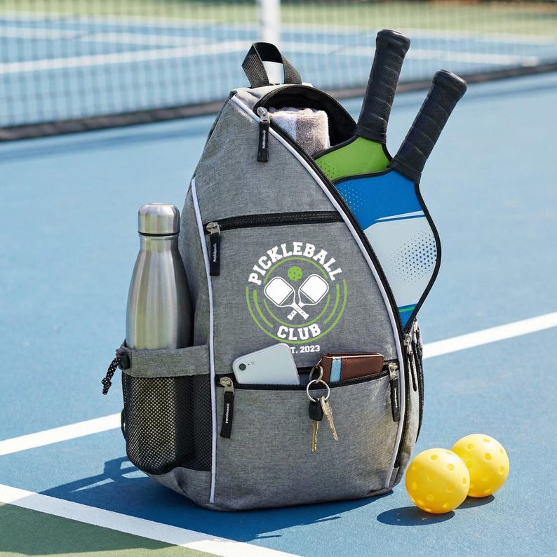 Adult pickleball backpack with a custom Pickleball Club logo, containing a water bottle, two tennis rackets, and two yellow pickle balls, is placed on the court.