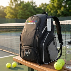 May include: A black backpack with a white Pickleball logo and colourful circular icons on the front. The bag features a side pocket holding a white paddle handle, a Clayco Sports logo on the lower front pocket, and mesh side panels with white trim.