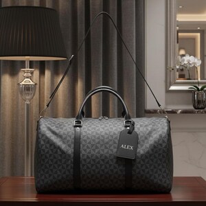 A gray patterned duffel bag with a black handle and tag is placed on a wooden table in a room with a lamp and a mirror.